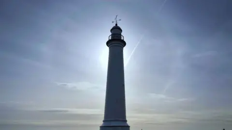 Mackemlan A tall white lighthouse with a metal wind vane on top. The lighthouse is largely in shadow and the sun in directly behind the structure casting a ring of light around it. The sky is a dusty blue colour and there are few clouds in the sky.
