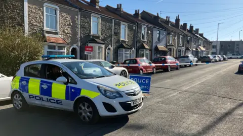 A police car parked across Filwood Road in Bristol. A blue police sign is on the floor and blue and white tape is cordoning off the street. Houses and parked cars can be seen in the background.