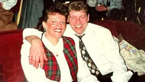 McCom's late family Ann McCombe and her husband Stanley were sitting together. There are curled black hair. She wore a white top and a grid. He has short brown hair, wearing a white shirt and spots. 