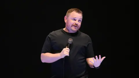 NI Comedy John Meagher stands, wearing a black t-shirt, alone on stage, holding a microphone 