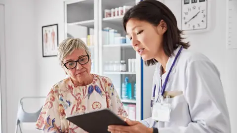 A brown haired Female doctor sharing electronic test results with light haired female patient at a clinic. Medical expert and woman are examining reports. They are sitting in examination room.