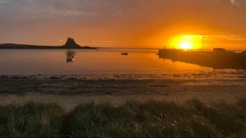 Sarah Hills The sun is setting beyond a fishing pier with Lindisfarne Castle rising like a volcano in the distance. The whole sky is bathed orange and the sea is completely calm with two small boats moored up beyond the shoreline.