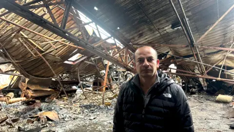 A man looking into the camera with a black north face coat on. In the background is the remains of a fire. The roof of the building behind him is falling in and there is clear ash and debris behind him as well. 