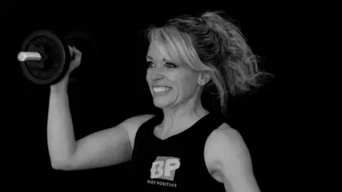 Sally Uphill Photography Loretta Davies pictured lifting a dumbell and smiling away from the camera. The photo is in black and white and she wears a "body positive" vest. 