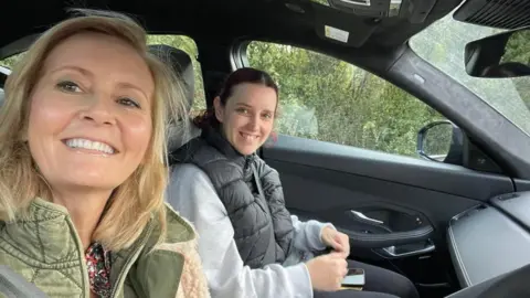 Contributed Two women sitting in the front of a car. They are both smiling and looking into the camera. One is blonde one and is brunette