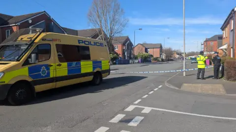 A police van is parked in the foreground with police tape cordoning off a street behind it. Two officers stand by the cordon talking. Another police van can be seen in the distance at the other end of the cordon.