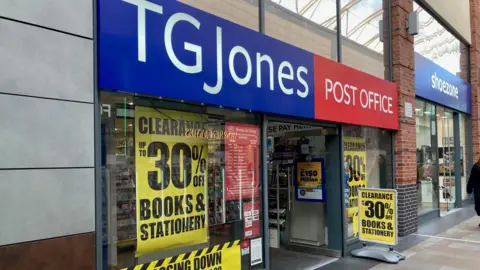 LDRS The exterior of a retail store named TG Jones, which also functions as a Post Office. The store has a large blue and red sign above the entrance, with “TG Jones” in white letters on a blue background and “Post Office” in white letters on a red background. The shop windows display yellow and black clearance signs.
