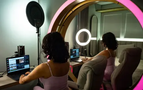 Jorge Calle / BBC The back of a woman with dark shoulder-length hair sitting at a desk looking at a laptop. She is wearing a light pink vest top. Her reflection can be seen in a large circular mirror, which has a bright pink frame. A bed can also be seen in the mirror.