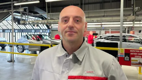 Andy Watson/BBC Jamie Whillians. He is standing in front of a production line of cars in the Nissan factory at Sunderland. His upper half is visible, and he is wearing a grey button up, with a Nissan logo on his left-hand side.