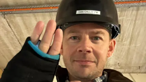 Dan Cheetham A man wearing a black hard hat holds up three fingers. His hand is in what appears to be a black cast.