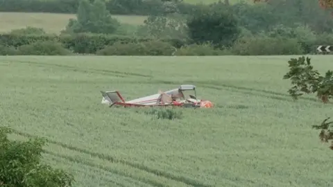 A light aircraft is seen in a field. It is mainly white with some red stripes on it.