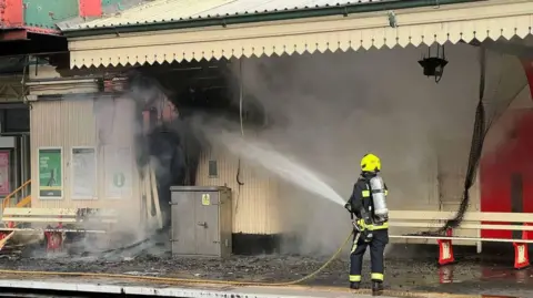Devon and Somerset Fire and Rescue Service The image shows a firefighter standing on a railway platform directing a high-pressure hose into a heavily damaged station building. Thick grey smoke is billowing from the structure, parts of the wall and roof appear blackened by fire, and debris is scattered across the wet platform surface in front of the building.

