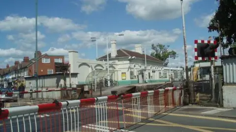 Portslade level crossing, protected by red and white gates, at Portslade station, which has two platforms and a white and green building