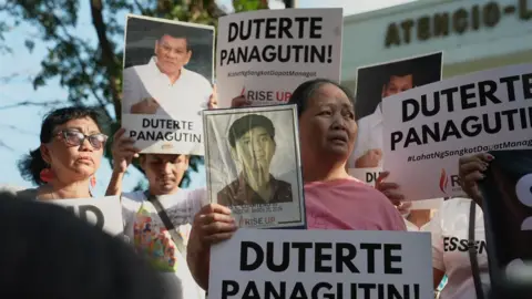 Virma Simonette Rivera/BBC News A group of people stand outside a building holding placards, some of them with pictures of Rodrigo Duterte, some with a picture of a young man, and others saying "Duterte panagutin", meaning "Duterte take responsibility"