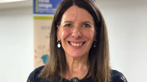 A woman with long brown hair is smiling widely and looking into the camera. 
