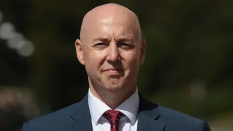A man, Jon Burrows, is staring at the camera. He is bald and is wearing a dark suit jacket over a white shirt and red tie. He has a neutral expression on his face. He is standing outside, the out-of-focus background appears to be trees. 