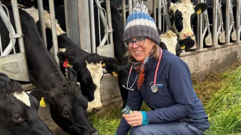 A vet crouches alongside some cows who are feeding on grass. She is dressed in a navy top and waterproof trousers with a colorful striped bobble hat. She has a stethoscope around her neck and a wide smile. 