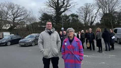 BBC Two shop owners stood in a car park. They are looking straight at the camera. One man is in a grey jumper. The lady is in a purple and pink coat. There are multiple people behind in the background along with some cars.