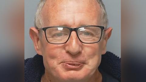 A police custody image of a smiling Neil Hutton, who is balding with short light grey hair and wears glasses and a blue fleece