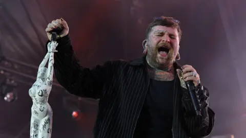 Chris Radburn/PA Wire A singer performs onstage. In his left hand is a microphone in which he is passionately singing, while his right hand rests on a microphone stand.