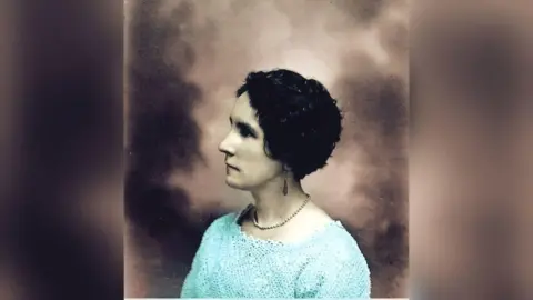 Gabrielle Deans A woman in a blue jumper, wearing a pearl necklace, looks to the side of the camera while posing in a photo studio. it is a old photogrpah.