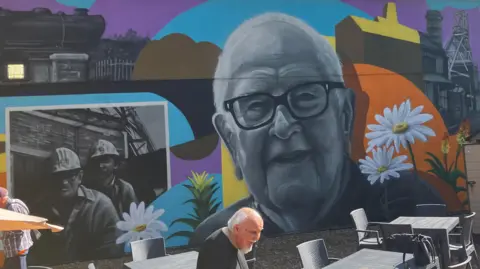 A mural featuring the greyscale image of a man's face surrounded by colourful representations of flowers and local landmarks