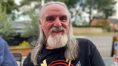 Pete Bevington wearing a blue t-shirt. He has long grey hair and a grey beard.