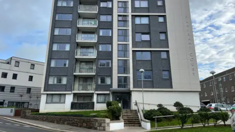 BBC The photo shows a tall, modern apartment building with multiple floors and balconies with a grey and white exterior. The building has large windows and a central entrance with steps leading up from the pavement. Vertically lettering spelling the name “Plaisant Court” is visible on the right side of the building.