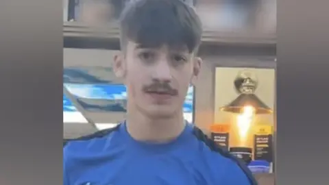 A picture of Taylor wearing a blue shirt with a white Nike logo on it. He has a short moustache and short hair.