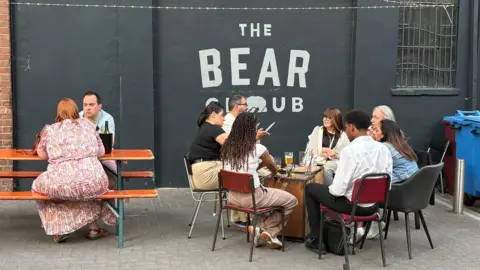 Martin Heath/BBC People sit at outdoor tables in a courtyard outside The Bear Club. A large black wall in the background features the venue’s name, “THE BEAR CLUB”, painted in white lettering with a bear logo. One group is seated around a low table with drinks, while two people sit together at a picnic bench. String lights hang overhead, and a brick wall, barred windows and recycling bins frame the venue’s outdoor seating area