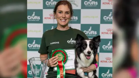 BeatMedia/The Kennel Club Laura, in a green T-shirt with hair tied back, has her arm around border collie Endeavour and holds a rosette in the other hand. A glass trophy is next to them.