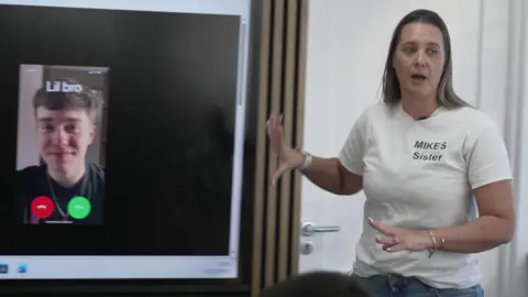 Becky Dustan, wearing a white T-shirt with "Mike's Sister" written on it talks to a classroom of pupils while giving a presentation about the effects of knife crime. On the interactive whiteboard is a screen shot from a phone showing a call coming in from someone called "Lil bro" with a picture of a young man showing.