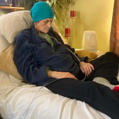Jared Wright Kelly lies on a hospital bed propped up by pillows and looking down, as if asleep. She wears a blue headscarf on and has a clear tube feeding into her nose. Her hands rest of her lap. 