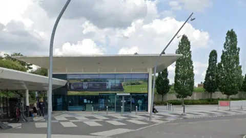 Google Corby station - a single-storey, glass-fronted building with a white roof. There is a white-roofed shelter to the left and trees to the right.