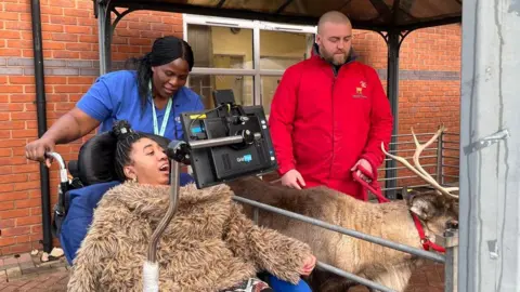 Alice Cullinane/BBC A woman is sitting in a wheelchair and using a screen to communicate. Her carer is standing behind the wheelchair as a reindeer stands next to them in a pen. 