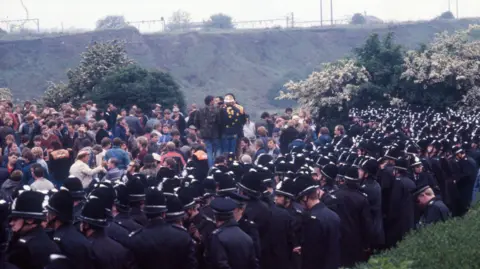 Another still from BBC footage. In front of a grass bank, two dense groups form facing each other - the protestors and the police officers.