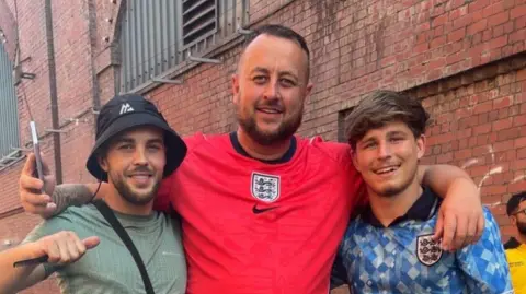 Family photograph Sean Carrington (centre) with friends Dwain Leonard and Rhys Cheetham wearing England shirts. They are Manchester's Depot Mayfield fanzone before the England v Argentina World Cup semi-final match was screened