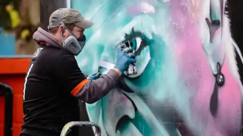 Jaime Bunting Photography David Nash, wearing a face mask, spraying his Keith Flint mural on to a water tank at Braintree Town FC.