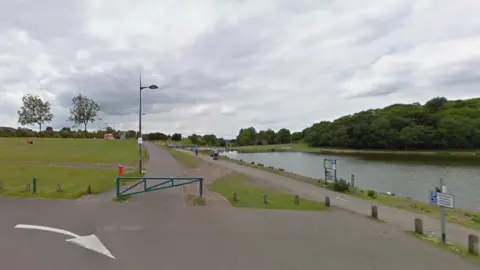 A view of a lake in a park next to a car park and path.