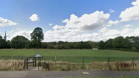 Google street view of the site in Wyndham Park in Grantham an open field with a black metal fence at the front and trees and a church steeple in the background