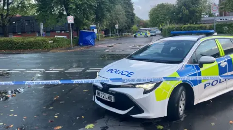 The scene of the stabbing with a police car in the foreground and a blue forensic tent in the background.
