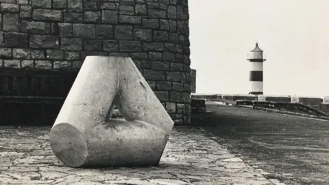 Portsmouth City Council A black and white image of a Portsmouth Marker. It is a stone structure in the shape of a triangle, with a brick wall in the background. To the right of the image is a lighthouse and a concrete wall lining the seafront.