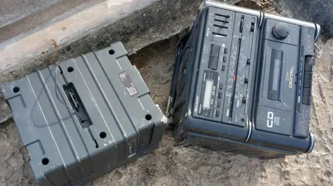 Wiltshire Council Image of an old, grey stereo system which has been dumped in the mud. In the background can be seen sheets of asbestos.