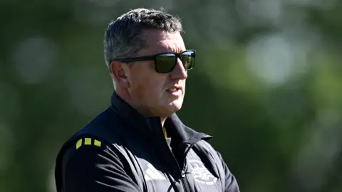 Gloucestershire director of cricket Jon Lewis stands wearing a black puffer jacket and sunglasses.