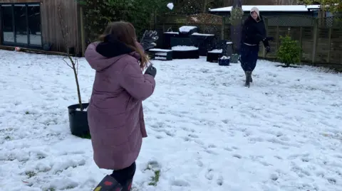 Sam Read/BBC Two children having a snow fight outside in a garden. There is snow on the ground, they are both wearing coats and gloves. There is a wooden building to the left and plants about.