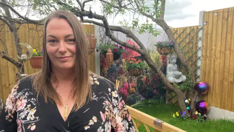 A woman with long, light brown hair and wearing a black top with flowers on it, is standing in her garden. The wooden boundary fence is behind her. In front of the fence is a tree, a buddha and other garden pots and ornaments.    