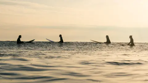 Carys Griffiths Laura Truelove and other female surfers sat on their boards in the sea as the sunsets and its orange glow reflects on the surface of the sea. 