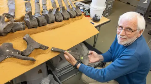 PA/James Brown Man with white hair and glasses wearing a blue jumper he is crouched next to a table with numerous dinosaur bones laid out.