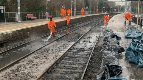 Greater Anglia Men in orange jackets cleaning up railway tracks with jets of water at a station. The tracks are covered in mud and there are bags on the platform.