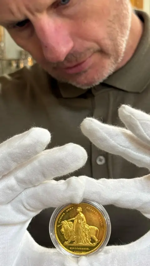 Rogers Jones and Co the coin in a clear case being held up between the index fingers and thumbs of a man wearing white gloves and a green shirt with his head down staring intently at the coin 